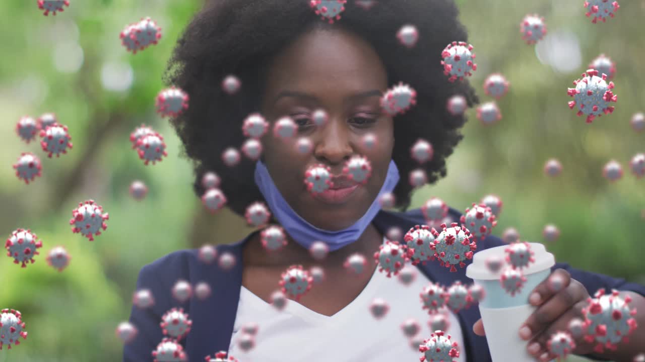 Animation of covid cells over happy african american woman with face mask drinking coffee in park