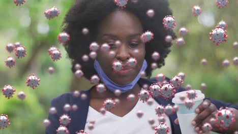 animación de células covid sobre una feliz mujer afroamericana con máscara facial bebiendo café en el parque