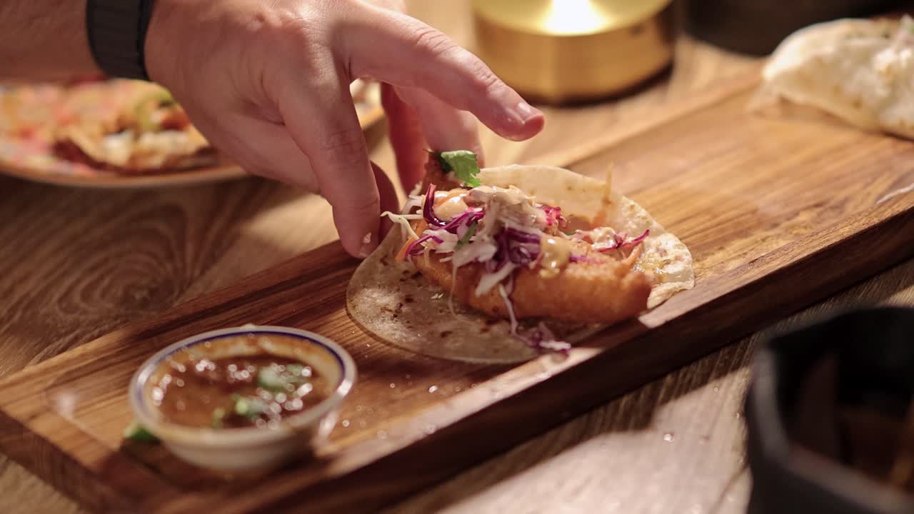 A hand reaches for and picks up a crispy fish taco topped with slaw from a wooden board, under warm indoor lighting, with shallow depth of field