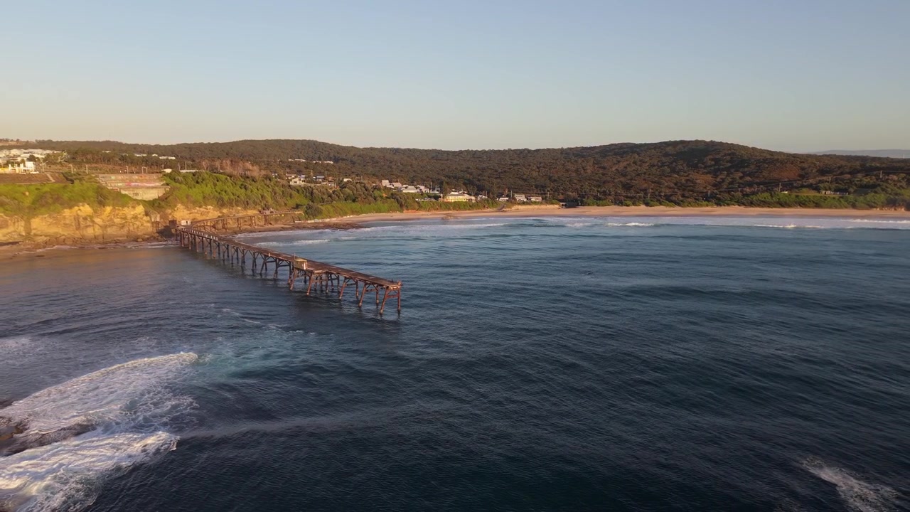 Cais de Catherine Hill Bay se estende da praia para a água costeira azul ao nascer do sol, com afastamento aéreo.
