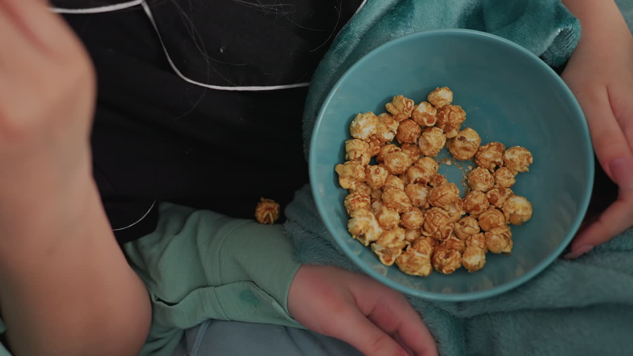 Close up of multiple hands reaching into bowl filled with caramel popcorn, suggesting shared snack moment during relaxed indoor activity