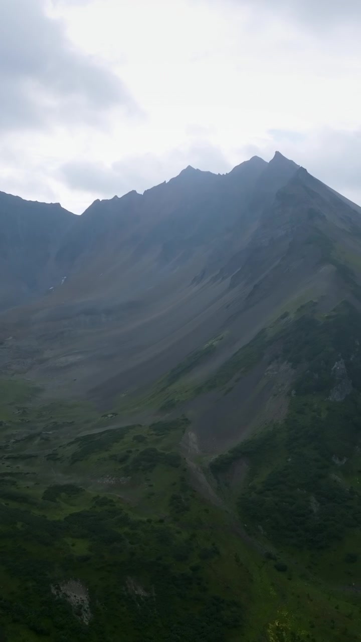 Rekaman udara yang luas dari pegunungan vulkanik Vachkazhets di Krai Kamchatka, Rusia, memperlihatkan puncak berbatu yang terjal, lembah pegunungan yang hijau, dan tundra di bawah langit musim panas yang mendung dan penuh awan