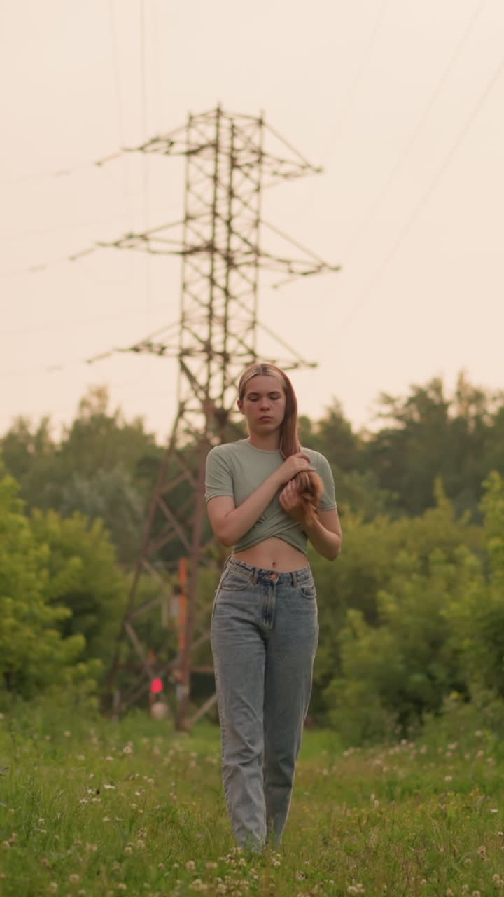 Young woman walking on grassy path, holding her hair forward with hands on her head, power lines stretching above, surrounded by lush greenery