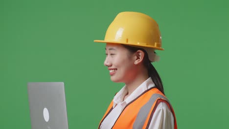 close up side view of asian female engineer with safety helmet working on a laptop and looking around while standing in the green screen background studio