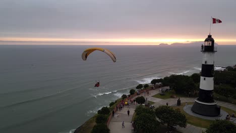 una toma aérea muestra un parapente volando al atardecer pasando junto a un faro y girando cuando las personas caminan en un bulevar debajo de él