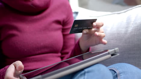 Midsection-of-biracial-woman-sitting-on-sofa-using-tablet-for-online-shopping-at-home,-slow-motion