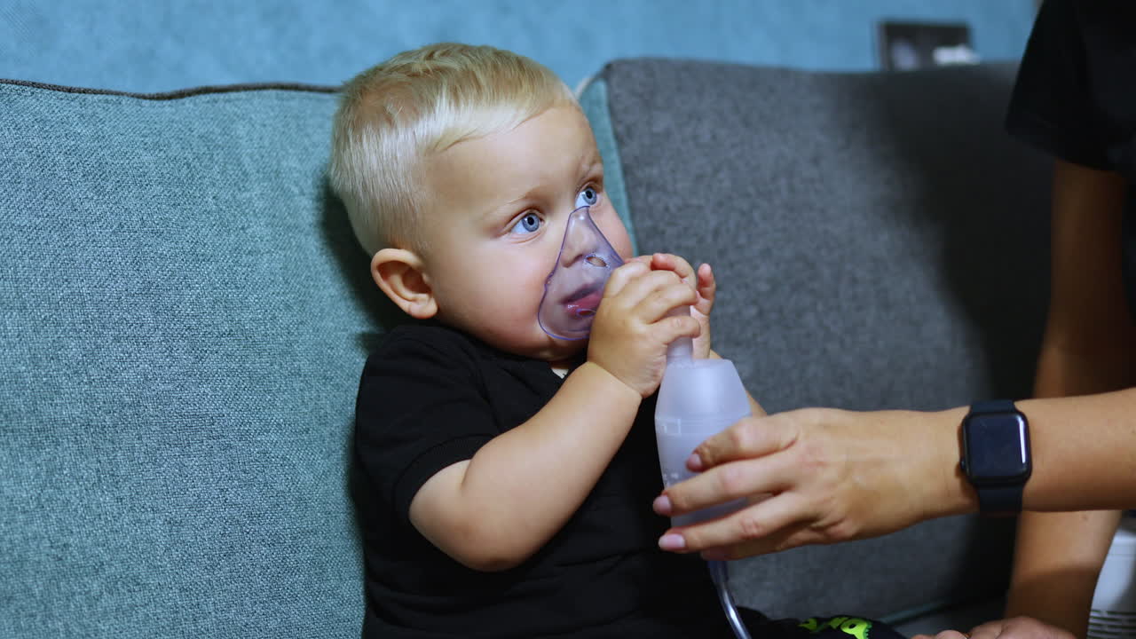 Un bambino piccolo è seduto sul divano e respira con un inalatore. Inalazione terapeutica per bambini. Maschera per inalazione per bambini
