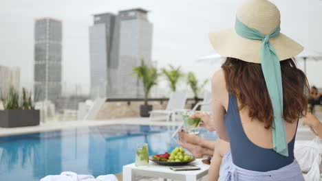 back view of gorgeous woman in stylish summer hat sitting next to a pool in a hotel building rooftop