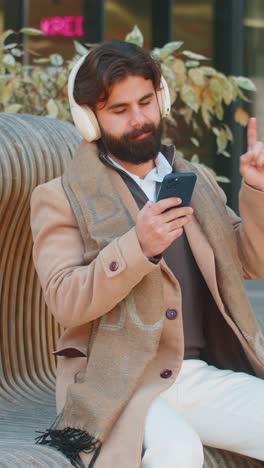 happy indian man in wireless headphones listening music in smartphone dancing sitting on bench