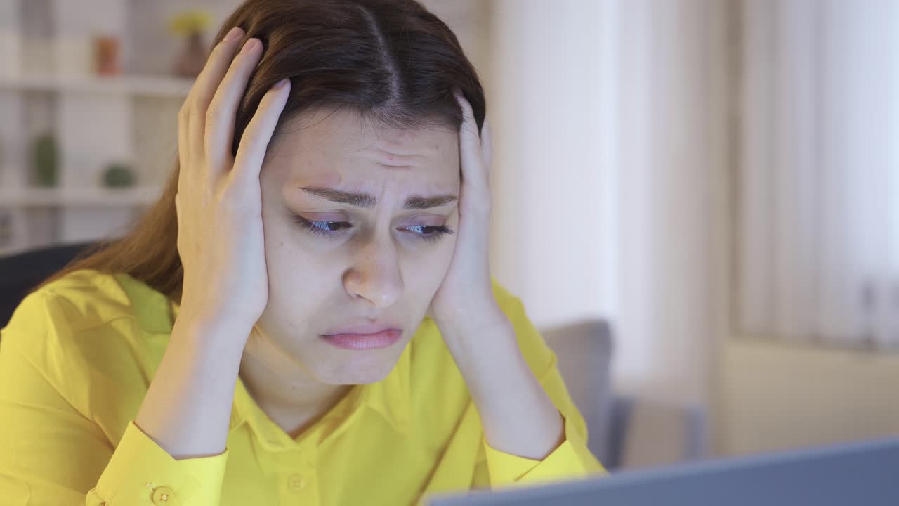 슬프고 걱정되는 젊은 여성이 집에서 노트북을 보고 좌절감을 느끼고 있습니다.