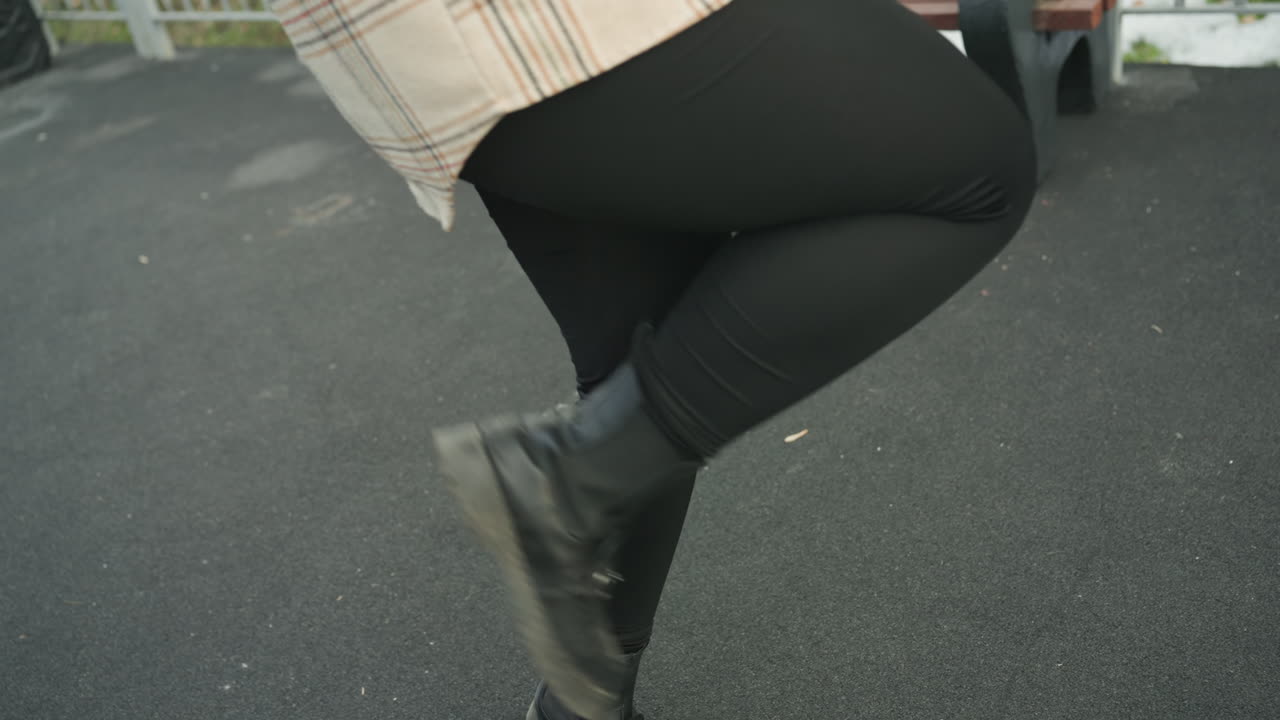 Street dance footwork on asphalt, closeup of woman legs in black leggings and worn boots, plaid coat hem visible, rhythmic stomps and precise steps, urban pavement texture and casual rehearsal energy