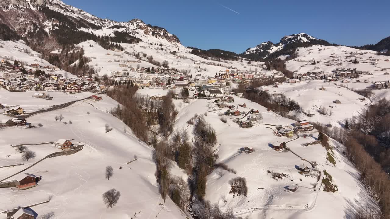 Kışın İsviçre Alpleri'ndeki Amden'e doğru çarpıcı drone yakınlaştırması; karlı zirveler, Alp köyü çatıları ve Walensee ile Glarus yakınlarındaki sakin dağ manzarası.
