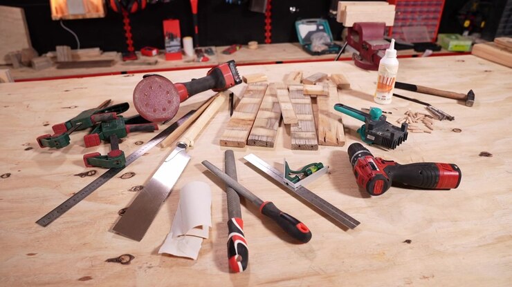 A collection of woodworking tools including a cordless drill, sander, hand saw, clamps and measuring tools lies beside timber pieces on a carpentry workbench.