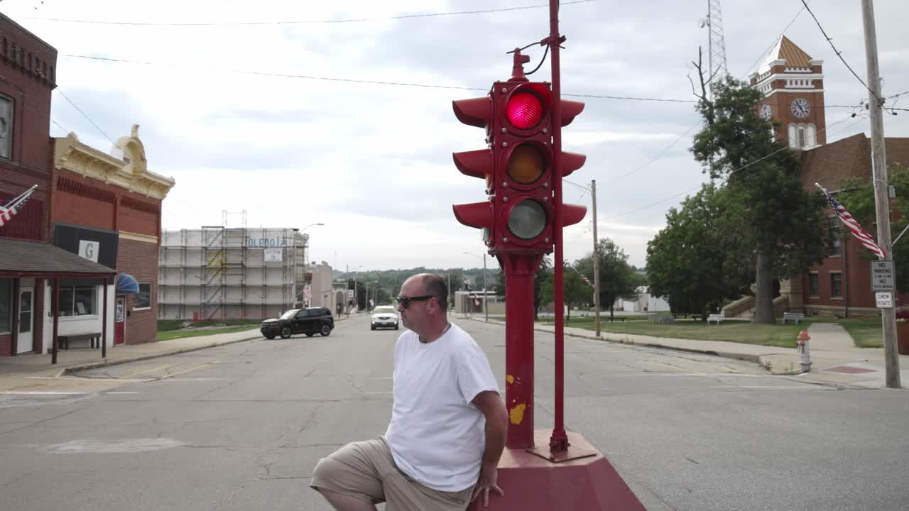 hombre con camisa blanca y gafas de sol sentado en la base de un antiguo semáforo de cuatro vías en el centro de toledo, iowa con video moviéndose alrededor