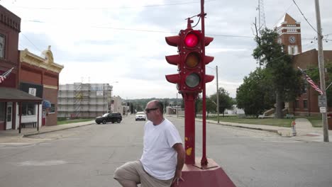 Hombre-Con-Camisa-Blanca-Y-Gafas-De-Sol-Sentado-En-La-Base-De-Un-Antiguo-Semáforo-De-Cuatro-Vías-En-El-Centro-De-Toledo,-Iowa,-Con-Un-Vídeo-Moviéndose