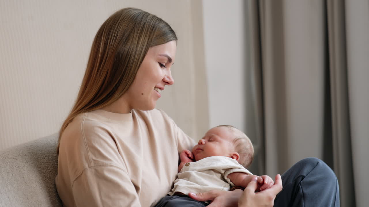 Happy smiling long-haired Caucasian woman waving her infant in arms sitting on a sofa. Mother strokes her child's arms with love.