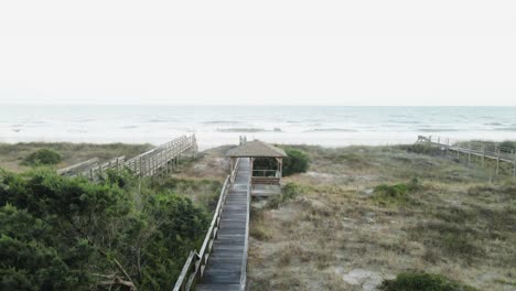 Auffliegen-Vom-Strandspaziergang-Mit-Pavillon-Und-Bäumen-Mit-Person,-Die-Am-Strand-Und-Dünen-Im-Hintergrund-Spazieren-Geht