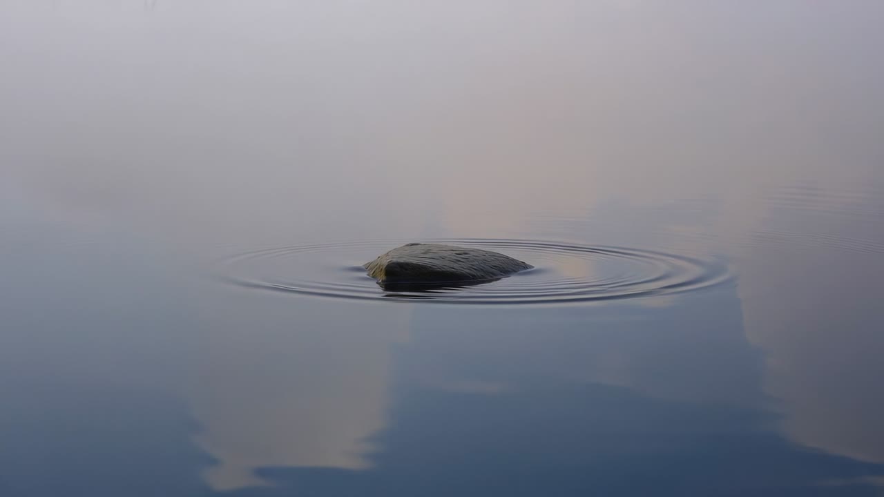 静かな池に広がる同心円状の波紋と空が映り込む、波立ち始めた暗い岩