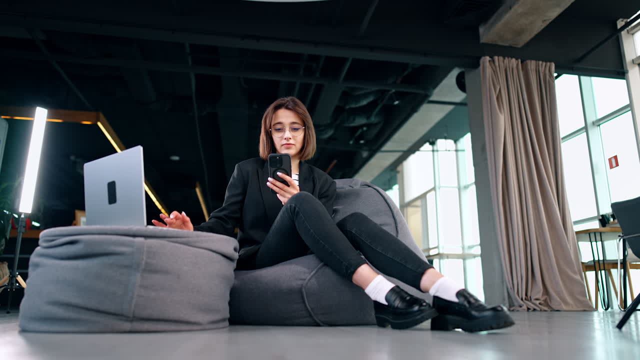 Attractive brunette woman sits in the bean bag chair near the ottoman with laptop on. Freelancing lady works in a comfortable space. Low angle view.