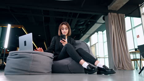 attractive brunette woman sits in the bean bag chair near the ottoman with laptop on. freelancing lady works in a comfortable space. low angle view.