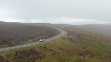 Oldtimer-Sportwagen,-Der-Auf-Nasser-Straße-Fährt,-Die-Sich-Zwischen-Wiesen-In-Der-Landschaft-Schlängelt.-Bewölkter-Tag-In-Dunstiger-Landschaft.-Irland