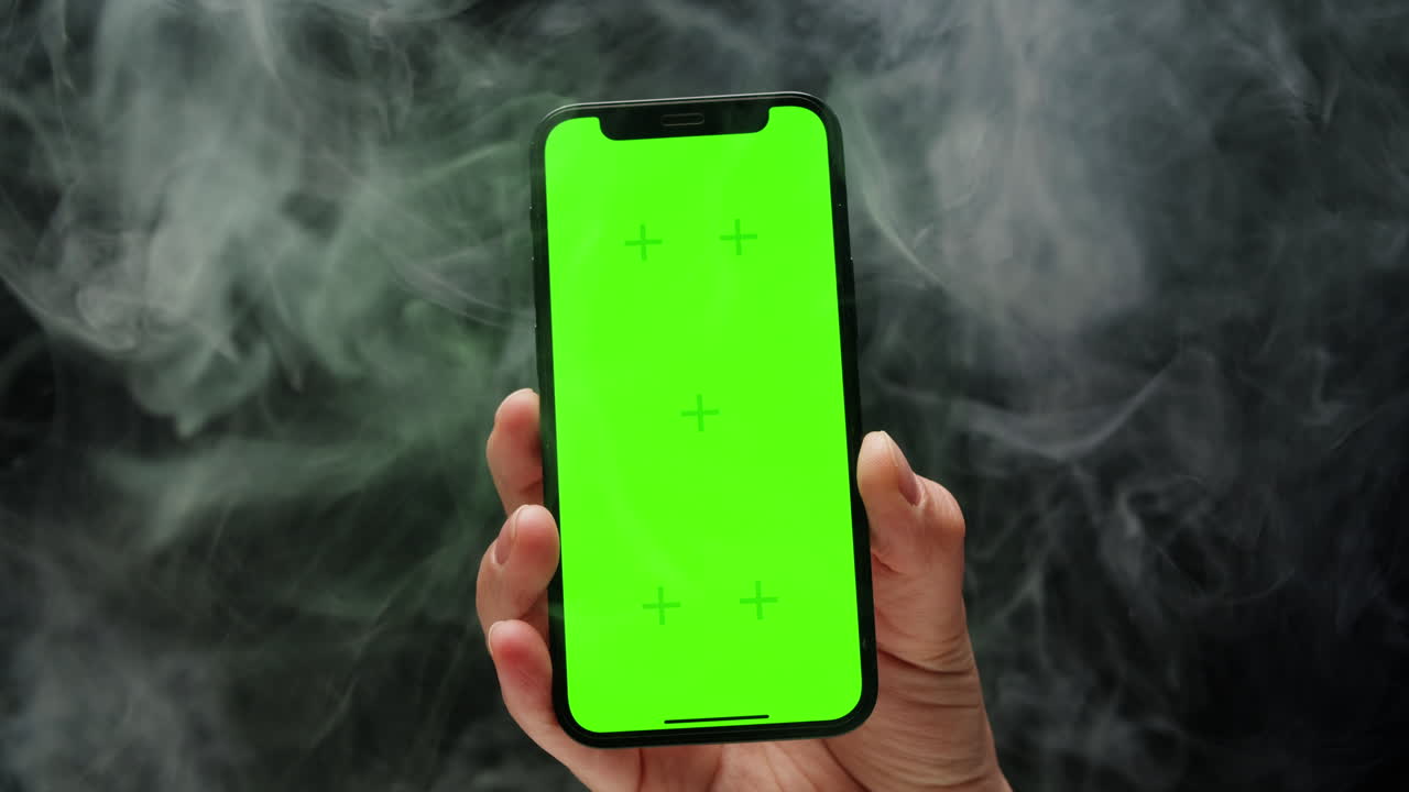 Smartphone with chroma key screen on black background in smoke close-up. Green screen on mobile phone in hand.