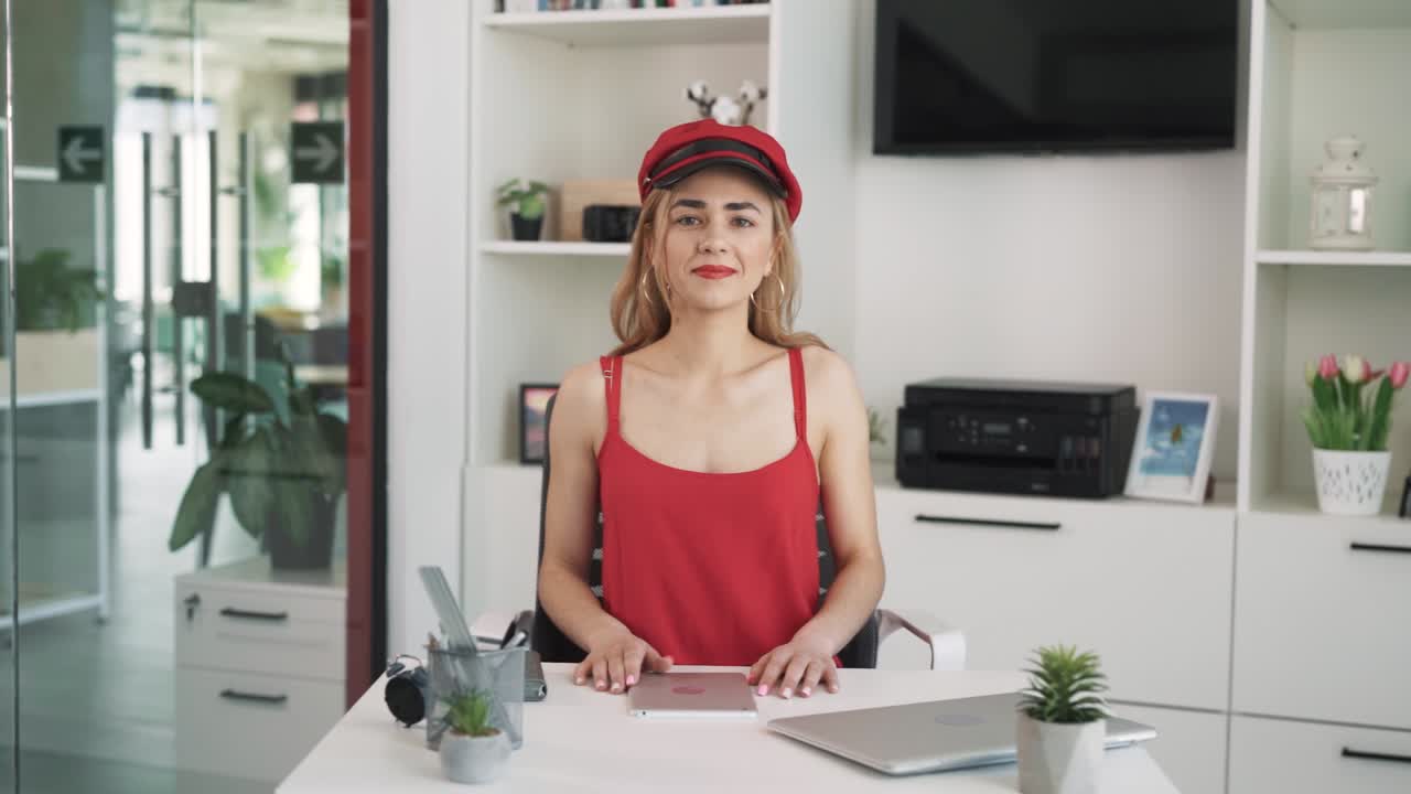 Young woman in a red dress and hat is sitting at a table in the office and looking seriously into the camera.A portrait of a young woman sitting at a table in the office.The office is modern