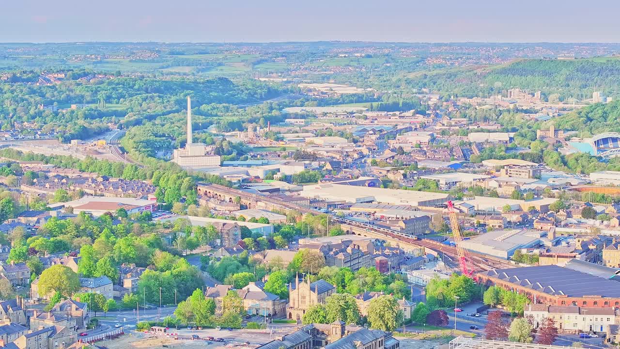 Expansive aerial view of Huddersfield from above Springwood, capturing warehouses, chimneys, rail lines, and dense greenery across the urban and semi-rural landscape, real time