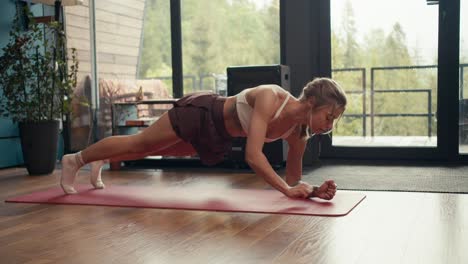 a blonde girl with tattoos in a white top does a dynamic plank exercise on a special mat in an industrial house overlooking a green forest. fitness at home, body and mind strengthening