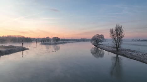 Toma-Panorámica-De-Un-Dron-Con-Un-Barco-De-Carga-Pasando-Por-El-Ijssel-En-Una-Mañana-Helada-Y-Brumosa-Con-Un-Hermoso-Amanecer