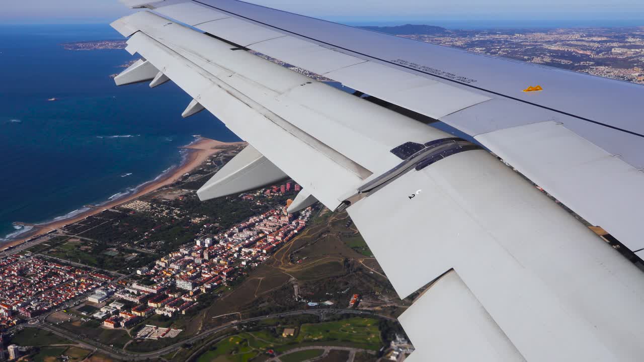 Point of view footage of a plane wing from inside a plane when it is flying over Lisbon sunny Portugal coastline and red rooftops at city while blue colored ocean is visible on the left with beaches