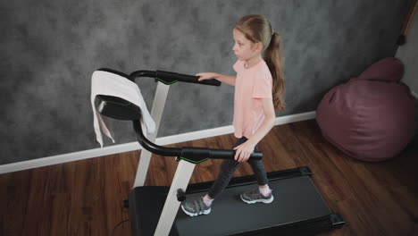 aerial view of young girl walking on treadmill holding handles wearing pink shirt patterned leggings in home workout room with grey textured wall wooden floor and seat visible