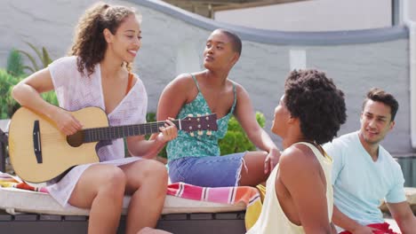 Glückliche-Biracial-Frau,-Die-Gitarre-Spielt-Und-Mit-Einer-Gruppe-Verschiedener-Freunde-Auf-Einer-Poolparty-Singt