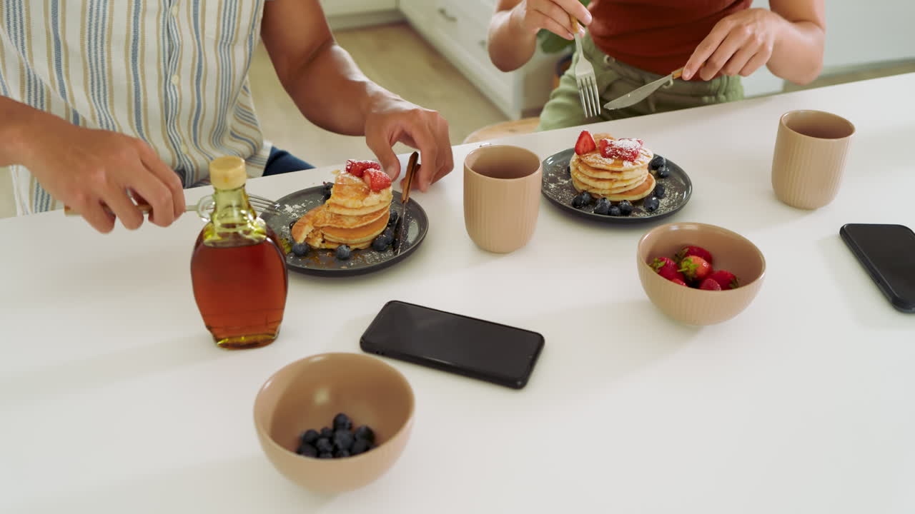 Diverse couple enjoying breakfast with pancakes and berries at home, sharing meal