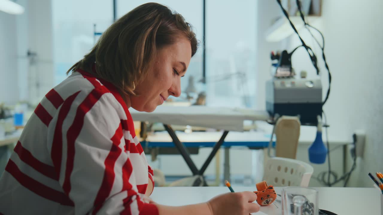 Focused woman sketches at a clean desk in a bright workshop. Her expression shows deep concentration on her creative task. She wears a red-and-white striped top while holding a pencil