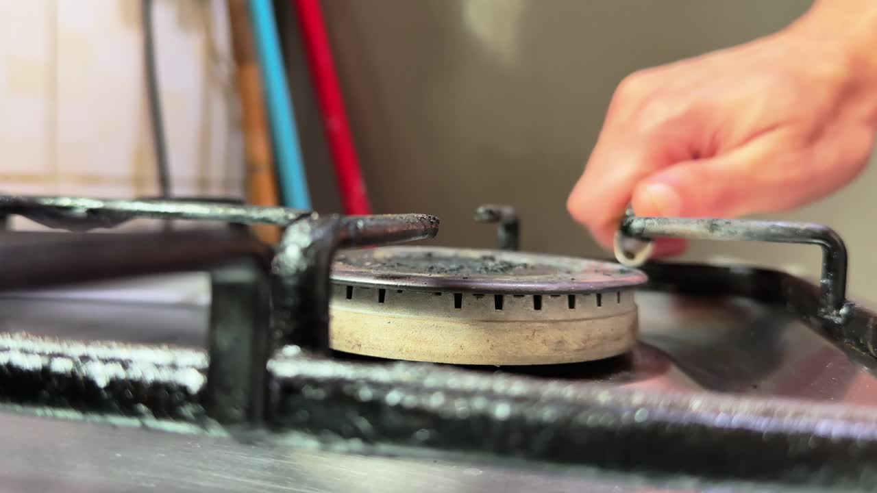 Close-up of a kitchen gas burner during ignition with a hand near the cooker, showing bright flame reflections on the metal stove in warm indoor light