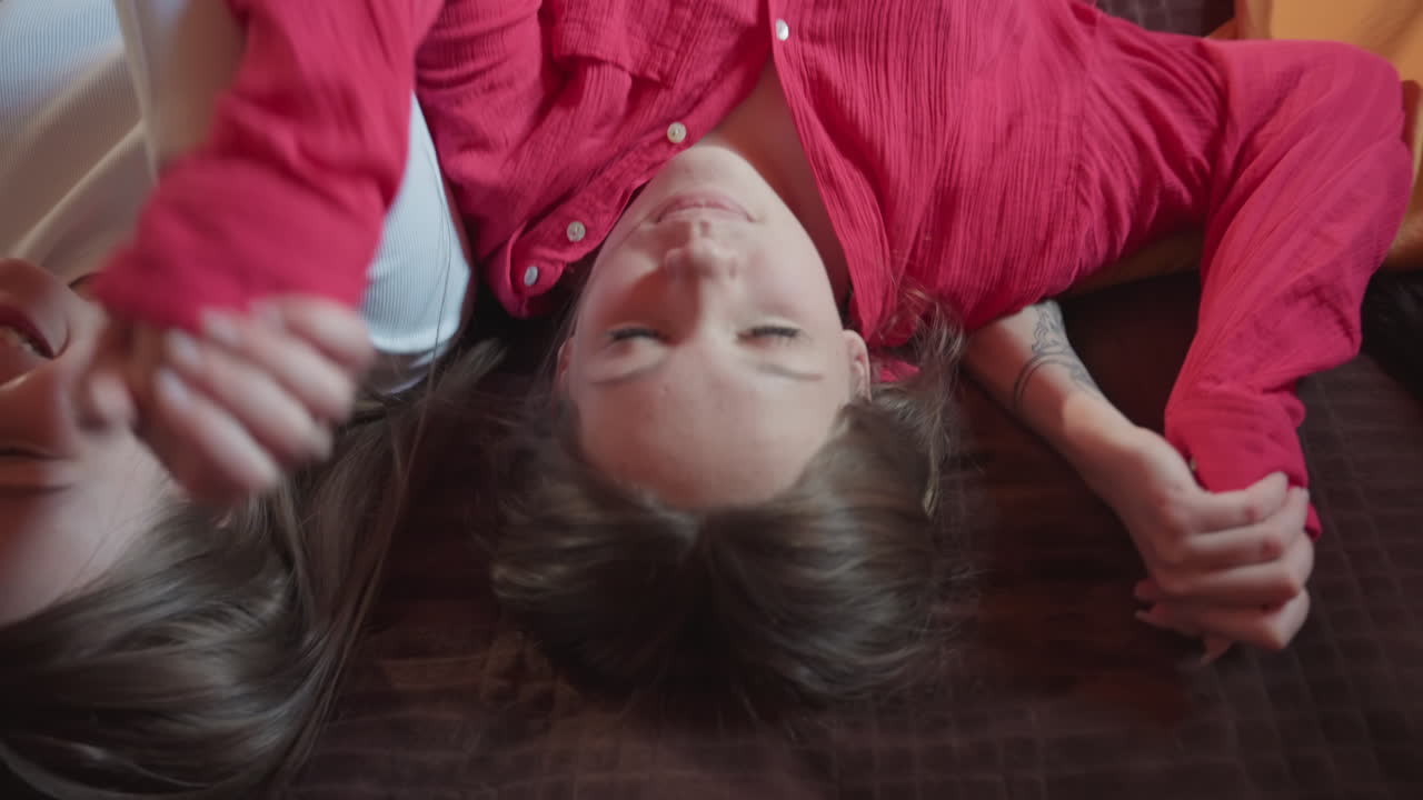 three young women preparing to rest backward on brown bedspread in warm wooden room, seen from behind, casual clothing and relaxed body language capturing mood of comfort