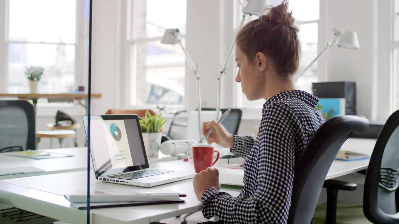 Young business woman working at shared desk in trendy hipster start up office using big data on laptop computer