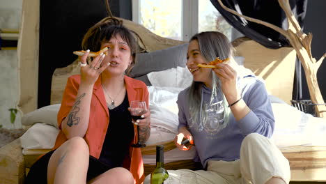 front view of two happy girls drinking wine and eating pizza while sitting on the floor and watching tv 1