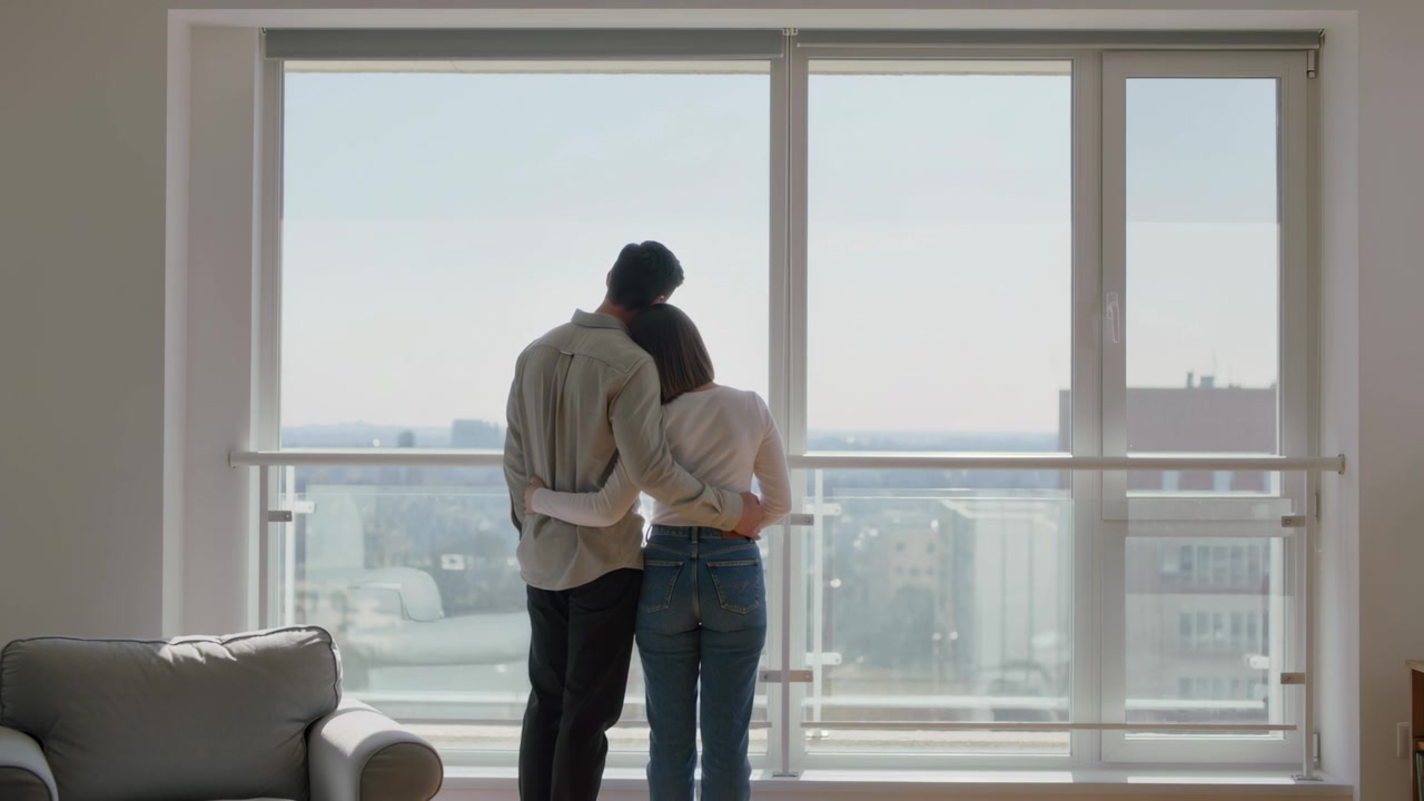 Afslappet par læner sig, nusser og kigger ud af stort vindue i lejlighed, hvor byens skyline trækker dem tættere sammen.