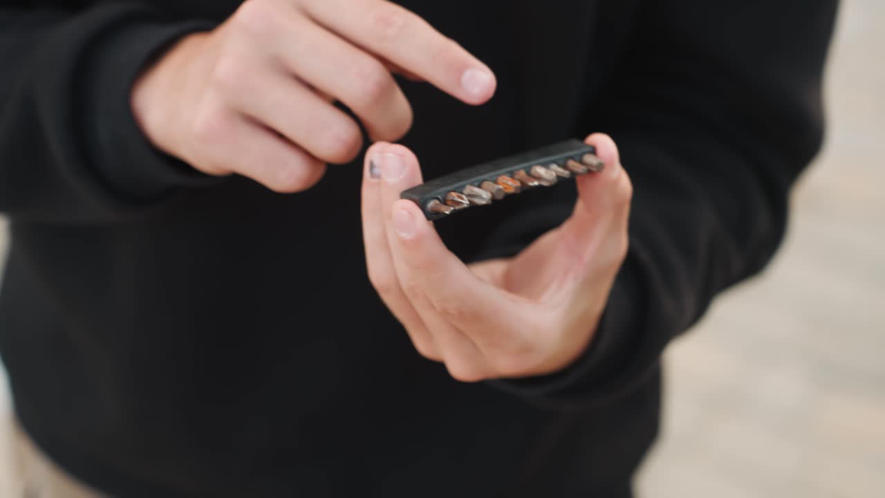 Close-up tangan memilih komponen logam di trotoar, nuansa DIY instruksional dengan hoodie dan posisi santai. Penjajaran ujung jari yang presisi pada komponen kecil, fokus dangkal dan latar belakang perkotaan, memuaskan.