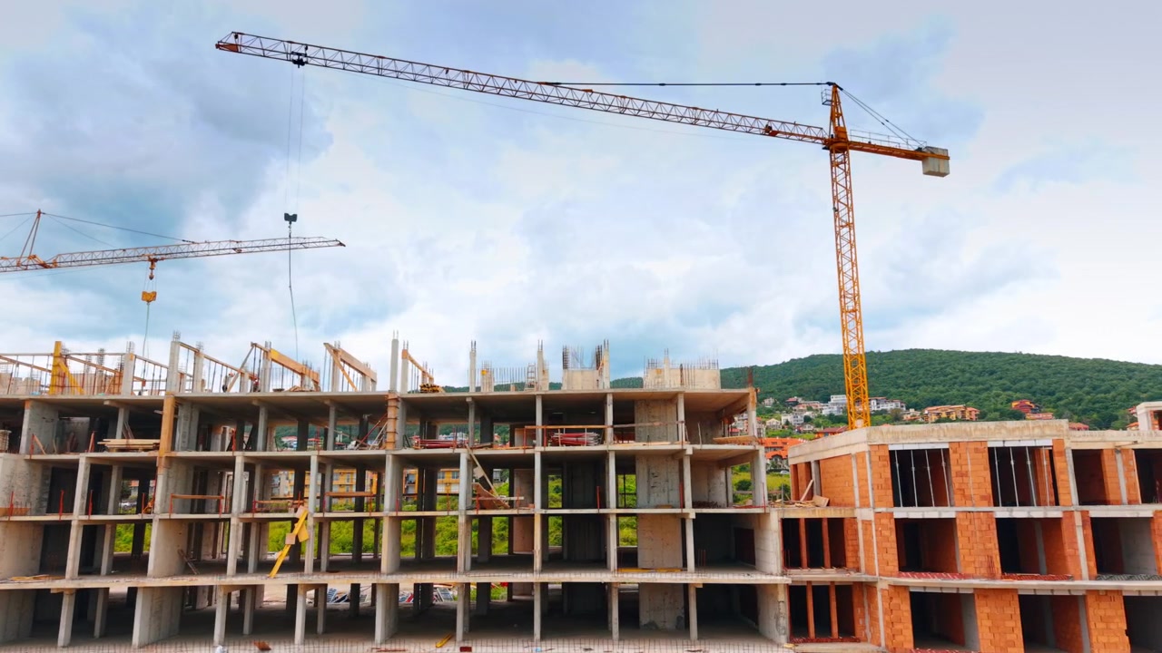Reinforced concrete building frame under construction with tower cranes. Straight on view of monolithic concrete structure skeleton with two yellow tower cranes against cloudy sky.