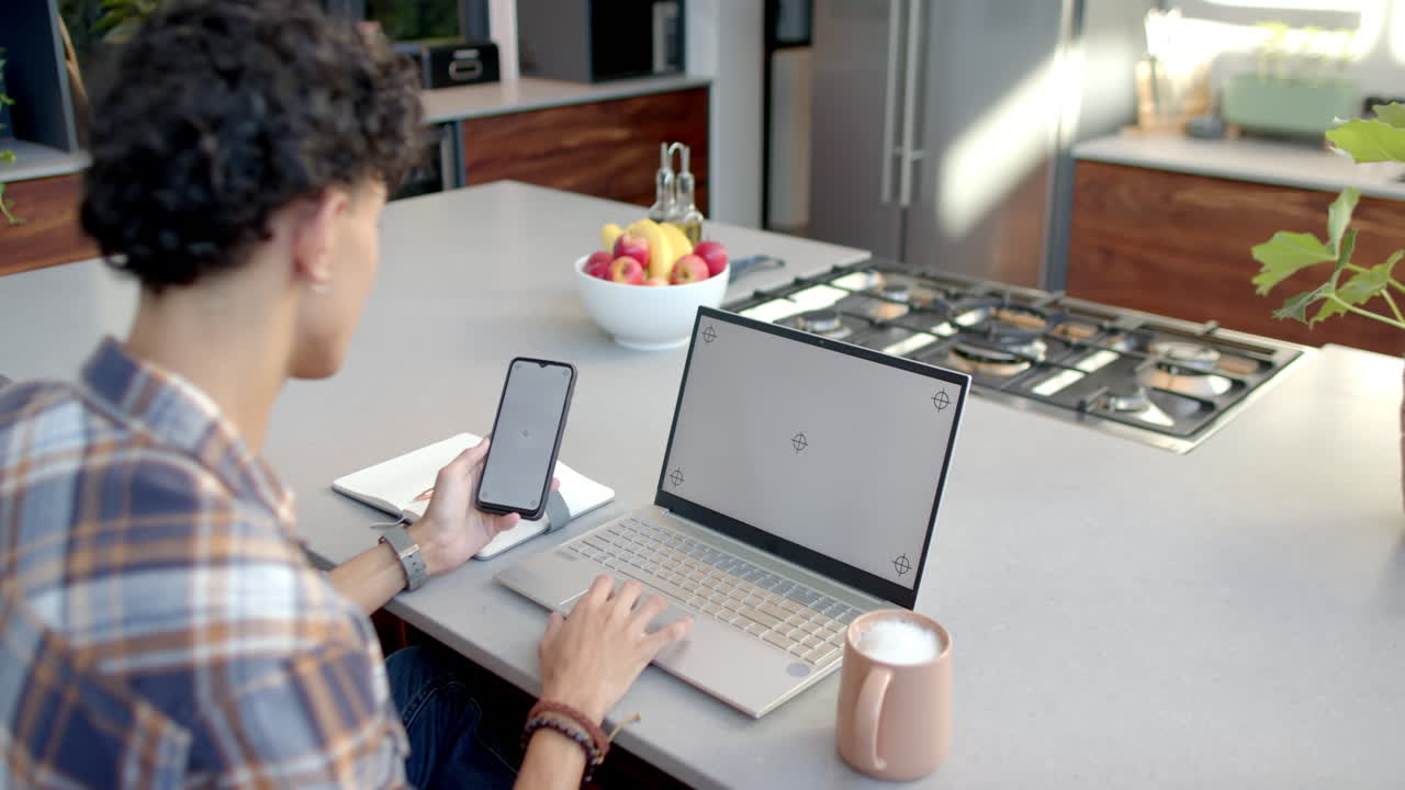 Using smartphone and laptop, teenage boy working from home in modern kitchen, copy space