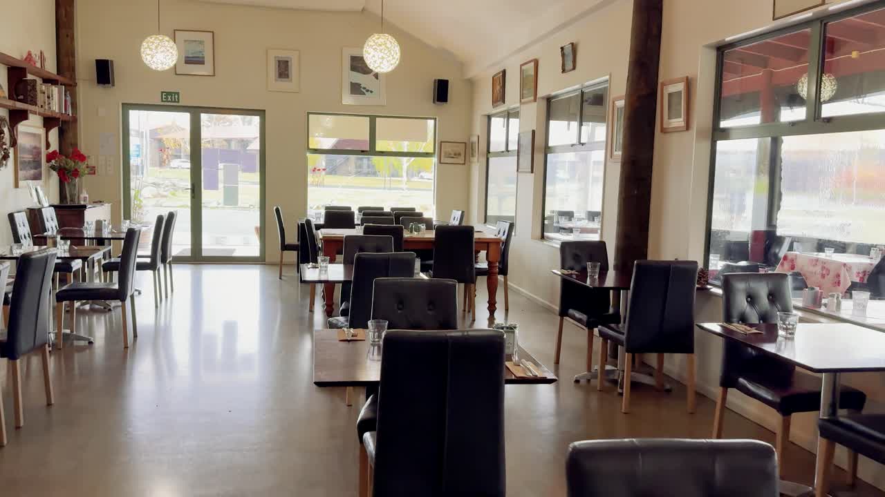 Camera slowly pans through a bright, contemporary cafe with empty tables and chairs, natural daylight streaming in, and a warm, inviting atmosphere