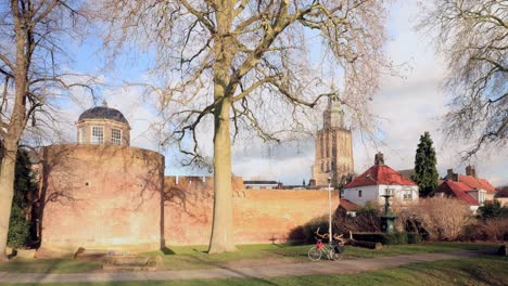 Zeitraffer-Der-Historischen-Stadtmauer-Mit-Park-Und-Festungsturm-Im-Winter-Karge-Landschaft-Mit-Walburgis-Kirchturm-Von-Zutphen,-Niederlande,-Im-Hintergrund-Vor-Blauem-Himmel-Mit-Wolken