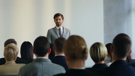 Caucasian-businessman-speaker-on-a-podium-wearing-formal-clothes-and-talking-in-a-conference-room-in-front-to-many-people