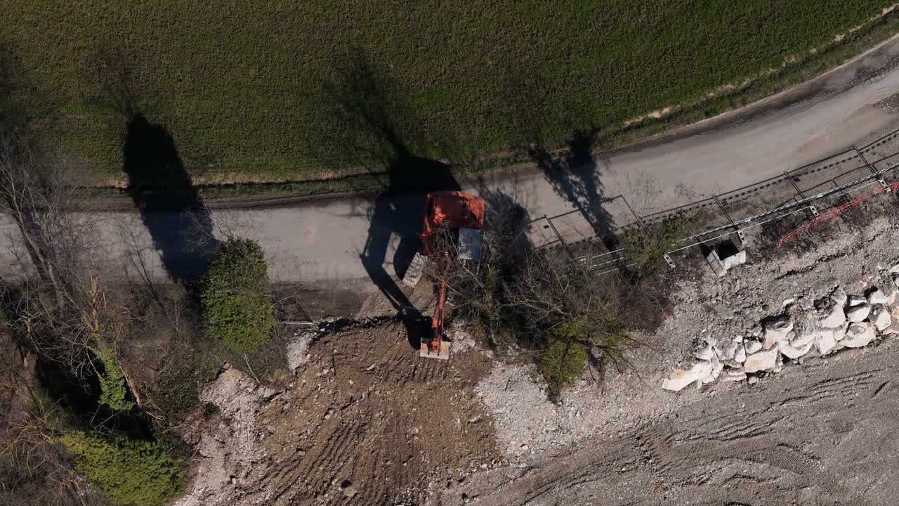 Drone top view of an excavator working beside a rural road, moving soil and rocks during construction or infrastructure maintenance operations