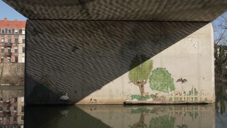 graffiti art at the wall near the reflective water surface, and concrete wall holding the bridge structure above displays an artsy message, location sarreguemines, france