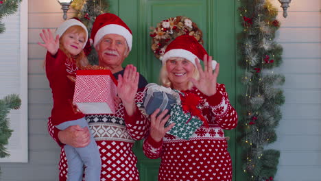 Senior-grandmother-grandfather-with-granddaughter-standing-at-Christmas-house-porch-waving-hello-hi