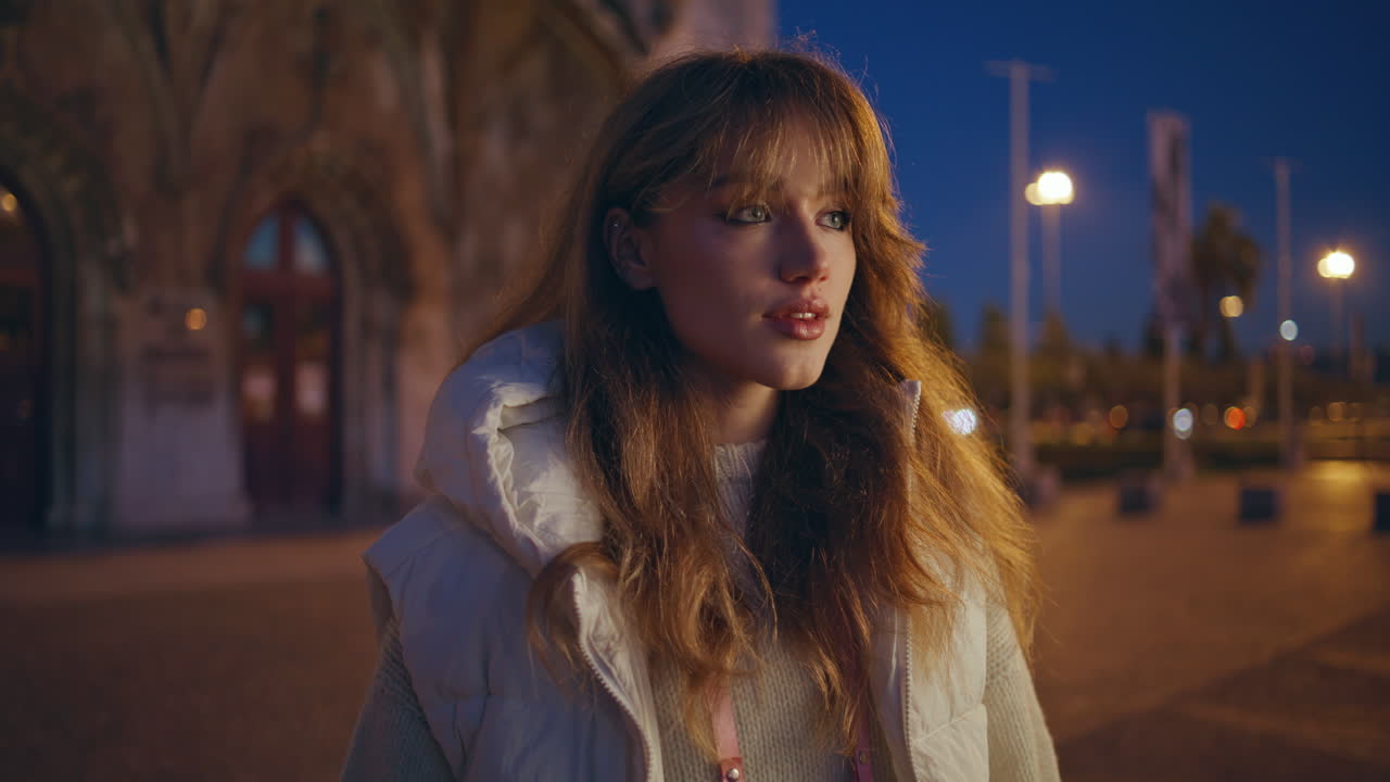 스타일리시한 여성이 저녁 시내에서 포즈를 취하고 머리카락을 조정하고 있습니다. 매력적인 갈색